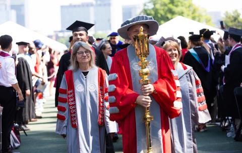 Image of Stephen Baker carrying the ceremonial mace during the academic procession. President Sally Kornbluth and 2023 speaker Mark Rober is seen behind Stephen Baker. 