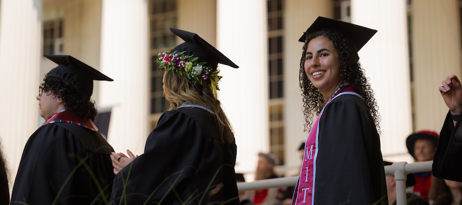 Image of a graduate waiting in line for her diploma and smiling out at the audience. The graduates seen in the photo are in their full regalia, cap and gown. The Building 10 columns are seen in the background.