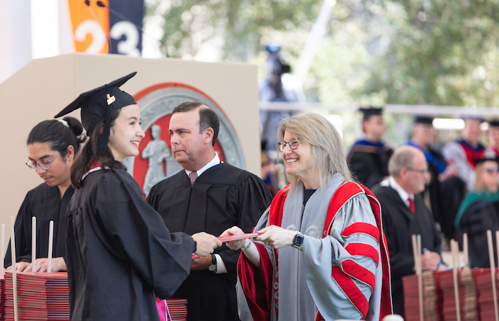 Image of President Sally Kornbluth smiling and handing a female student their diploma. 