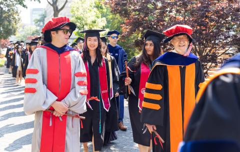 Image of the graduate division of the academic procession. Associate Marshals, Joel Voldman and Leslie Norford are seen in front of the students, with Class President Anna Sun and Undergraduate Association Vice President Shruti Ravikumar seen behind them. All are smiling