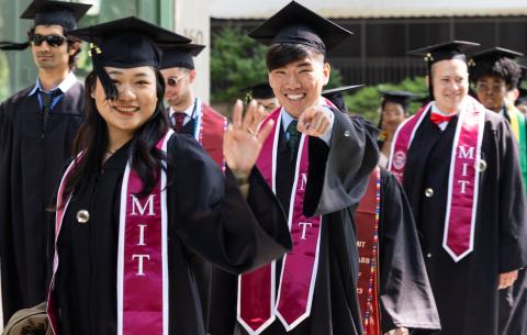 Image of graduates in their regalia, smiling and waving at the camera