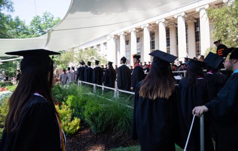 Image of undergraduates in line, walking onto the ceremony stage to receive their diplomas. The lobby 10 columns are seen in the background.