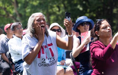 Image of a woman in the audience taking photos of the graduates on her phone. She is smiling with one hand raised to the side of her face.
