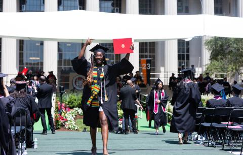 Image of graduate walking back to her seat after receiving her degree. She is smiling and one of her arms is raised in excitement.
